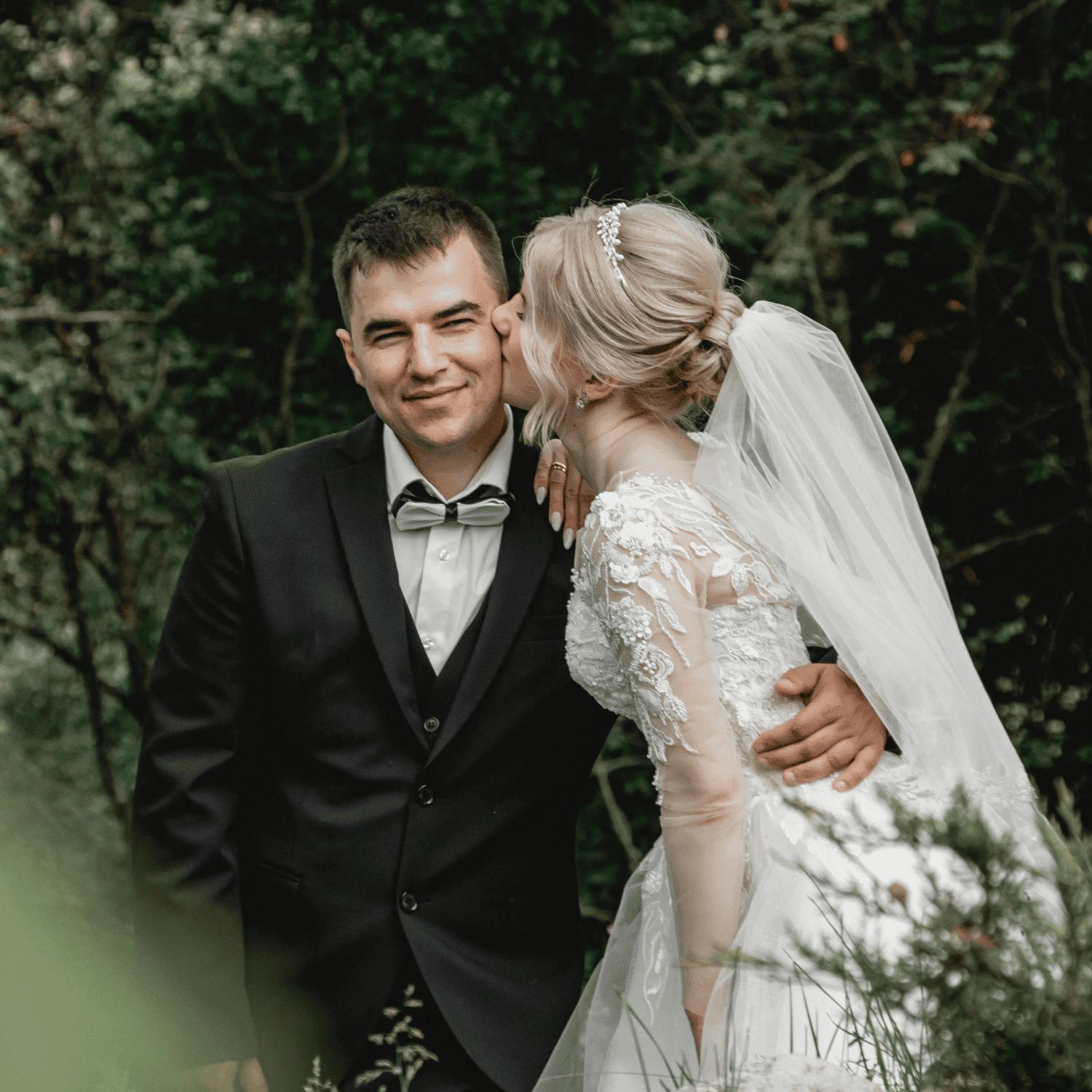Bride kisses groom's cheek in forest setting, both in elegant wedding attire.