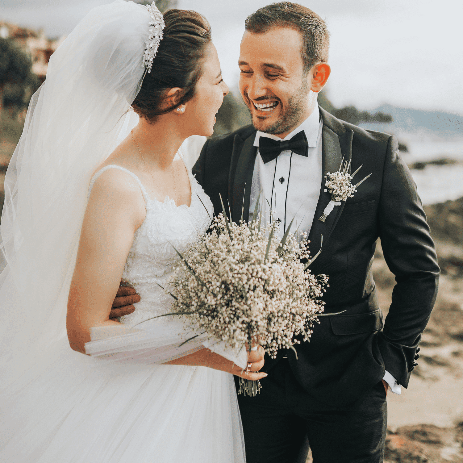 Bride and groom smiling, embracing on a rocky beach with flowers.