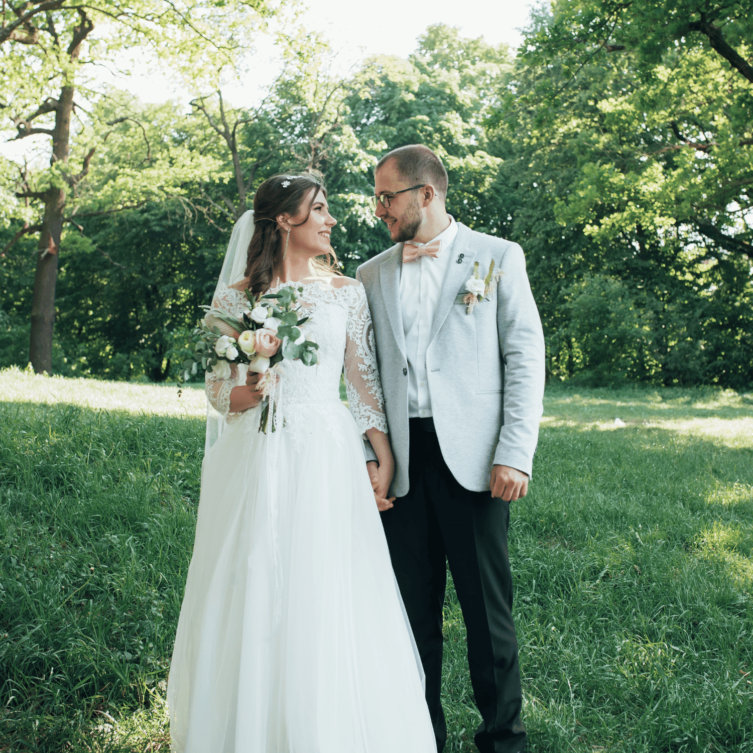 Bride and groom smiling in a park, holding hands, surrounded by lush greenery.